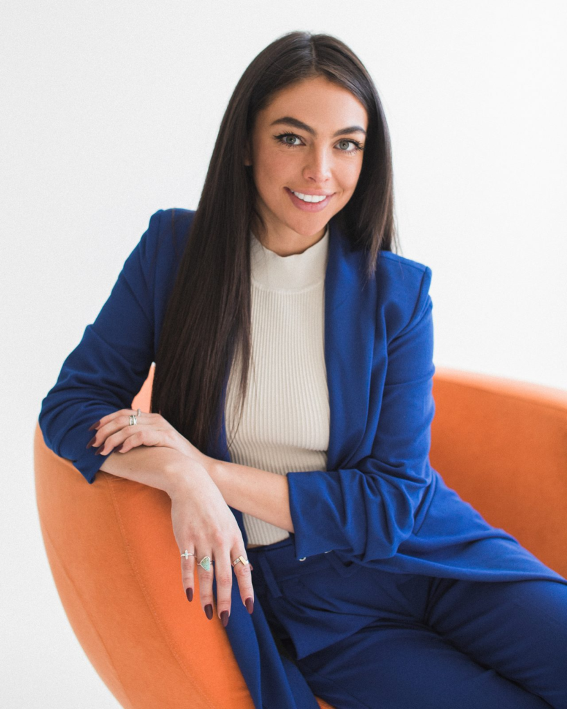 Sharissa Villanueva smiling, seated in an orange chair in a blue suit